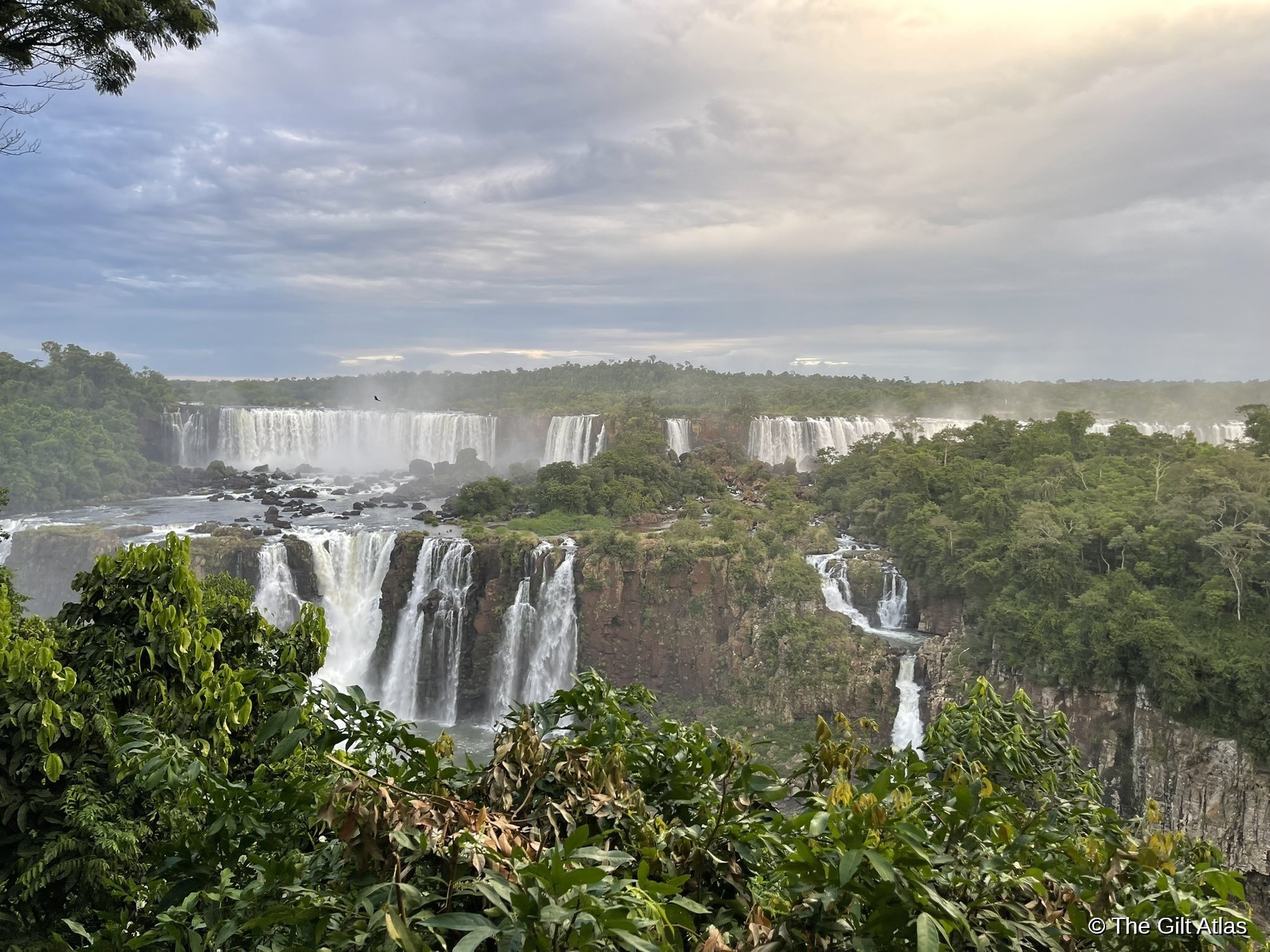 Iguazu Falls in sunlight — golden light breaking through clouds, full cascade visible with jungle in the foreground