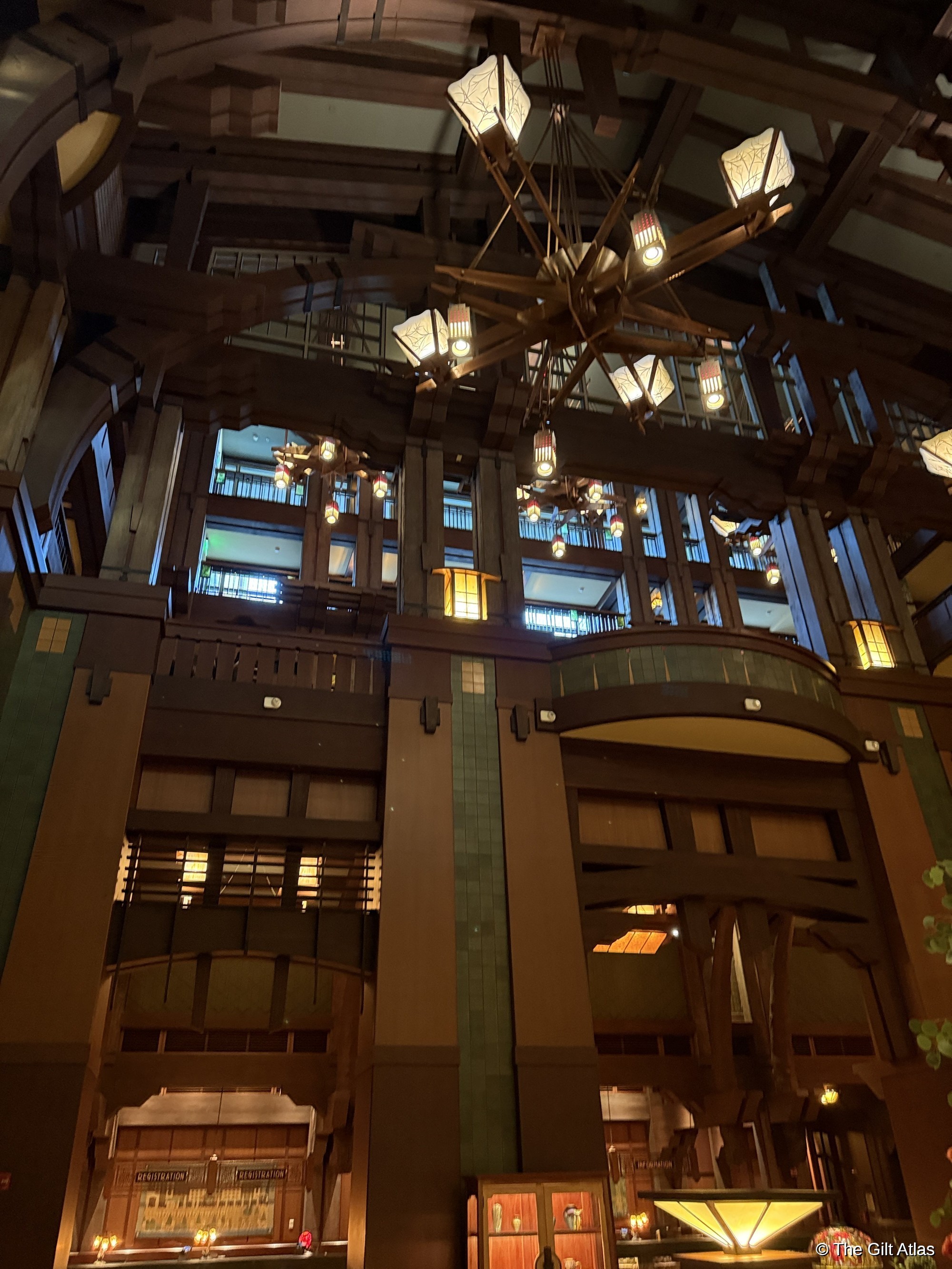 The Grand Californian atrium viewed from a balcony level &mdash; timber columns and beams, craftsman pendant lights, stacked balconies with green-tiled railings, and warm amber light throughout