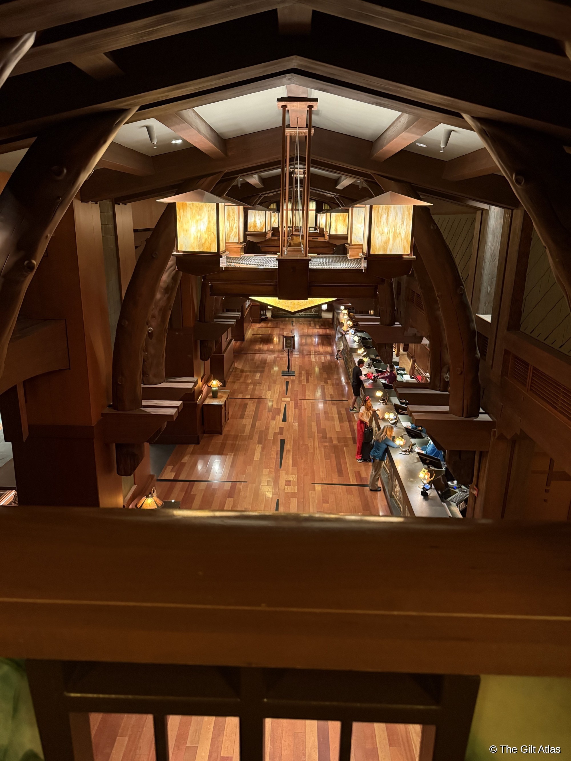 Looking down from a guest balcony into the Grand Californian Hotel &mdash; Arts and Crafts timber arches frame the view over the registration desk below, warm wood floors and craftsman pendant lamps visible along the corridor