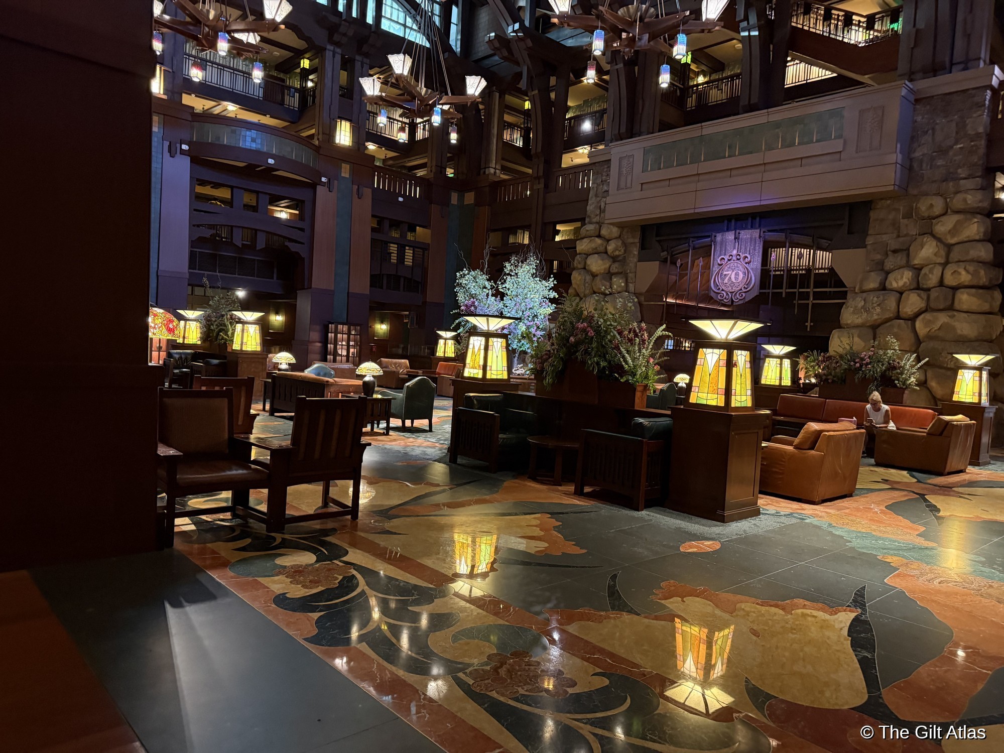 The Grand Lobby of the Grand Californian Hotel looking toward the main fireplace &mdash; stained glass table lamps, leather seating, Arts and Crafts floral carpet, and a towering floral arrangement on the central table, with the six-story atrium visible above