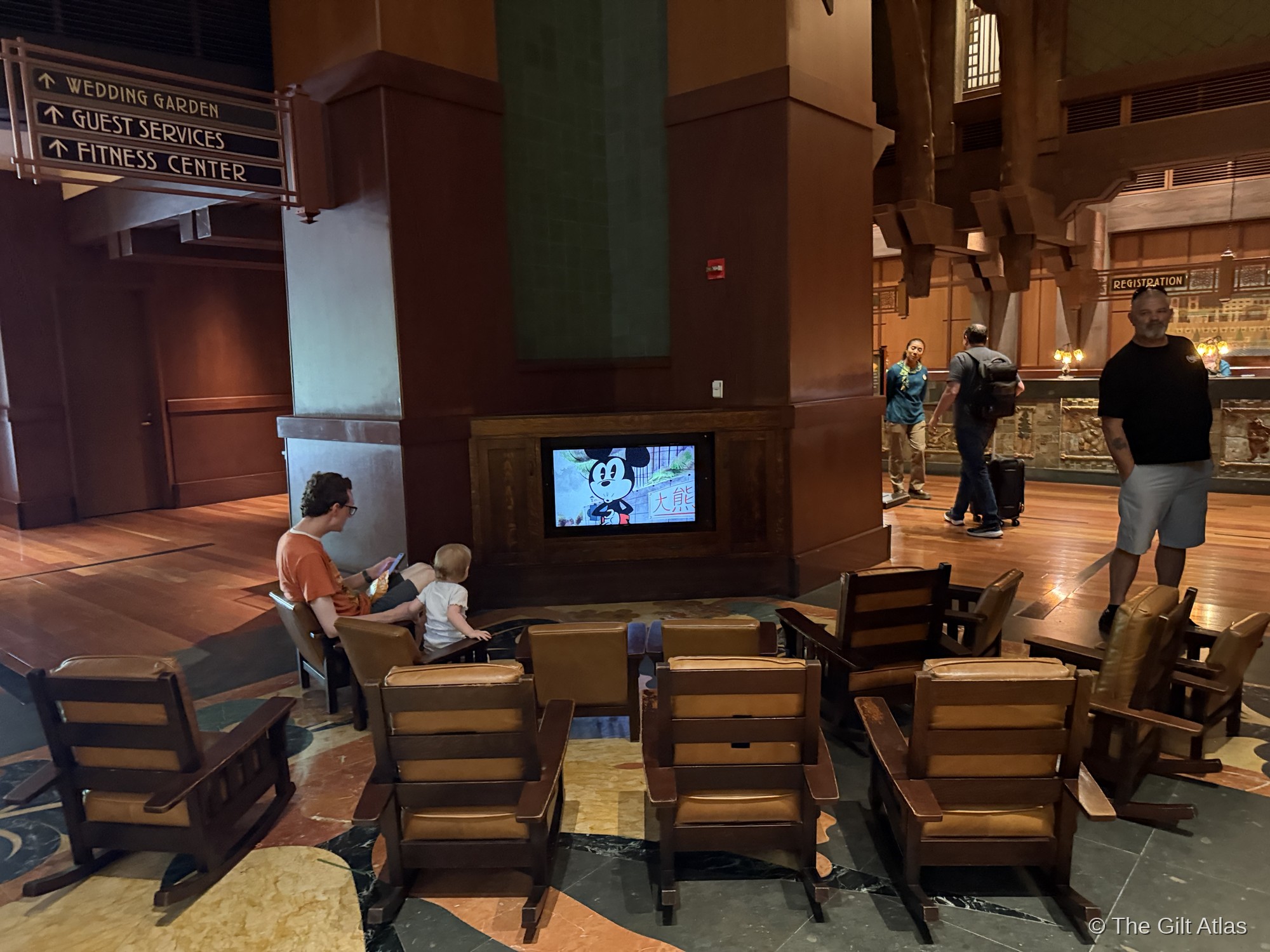 The Grand Californian Hotel lobby seating area &mdash; Mission-style rocking chairs arranged around a small Disney cartoons screen, with the Registration desk visible in the background and craftsman signage pointing to Guest Services and the Wedding Garden