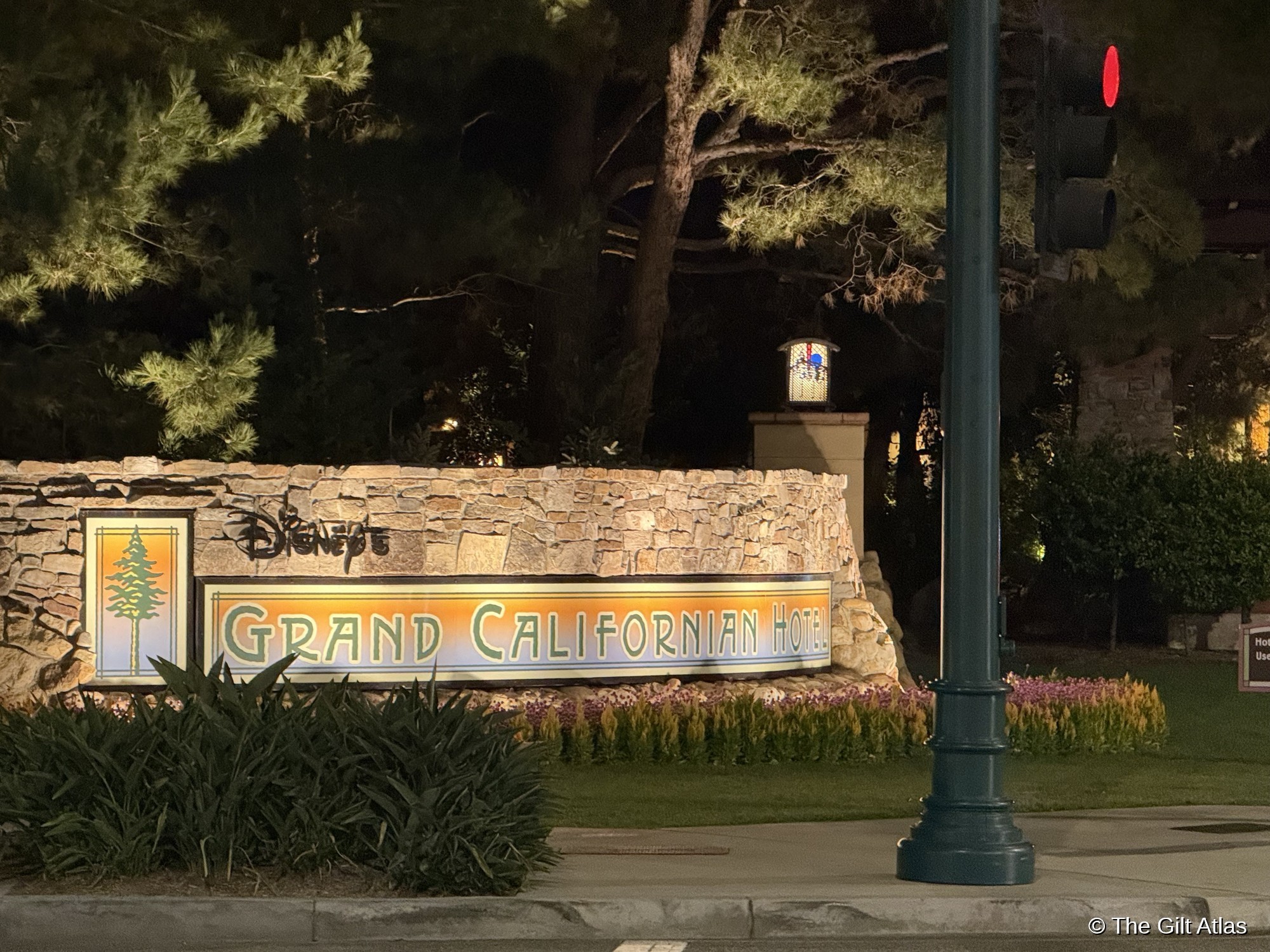 Disney's Grand Californian Hotel entrance sign at night &mdash; illuminated Arts and Crafts lettering set into a stone wall, with a craftsman lantern post and pine trees behind