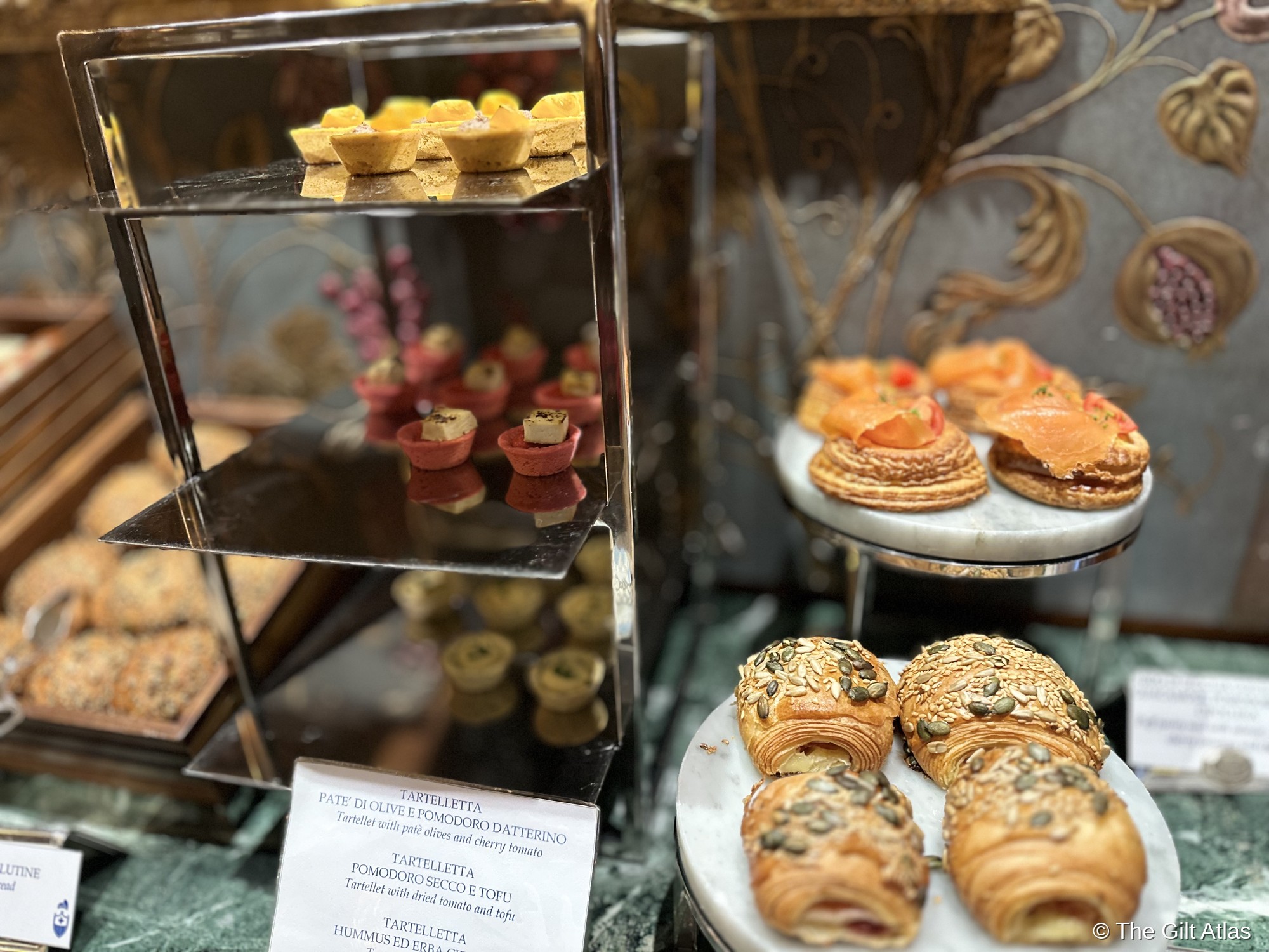 The Gritti Palace breakfast spread — savory pastries, smoked salmon, seed-topped croissants, and elaborate canapes against ornate gold Venetian wallpaper