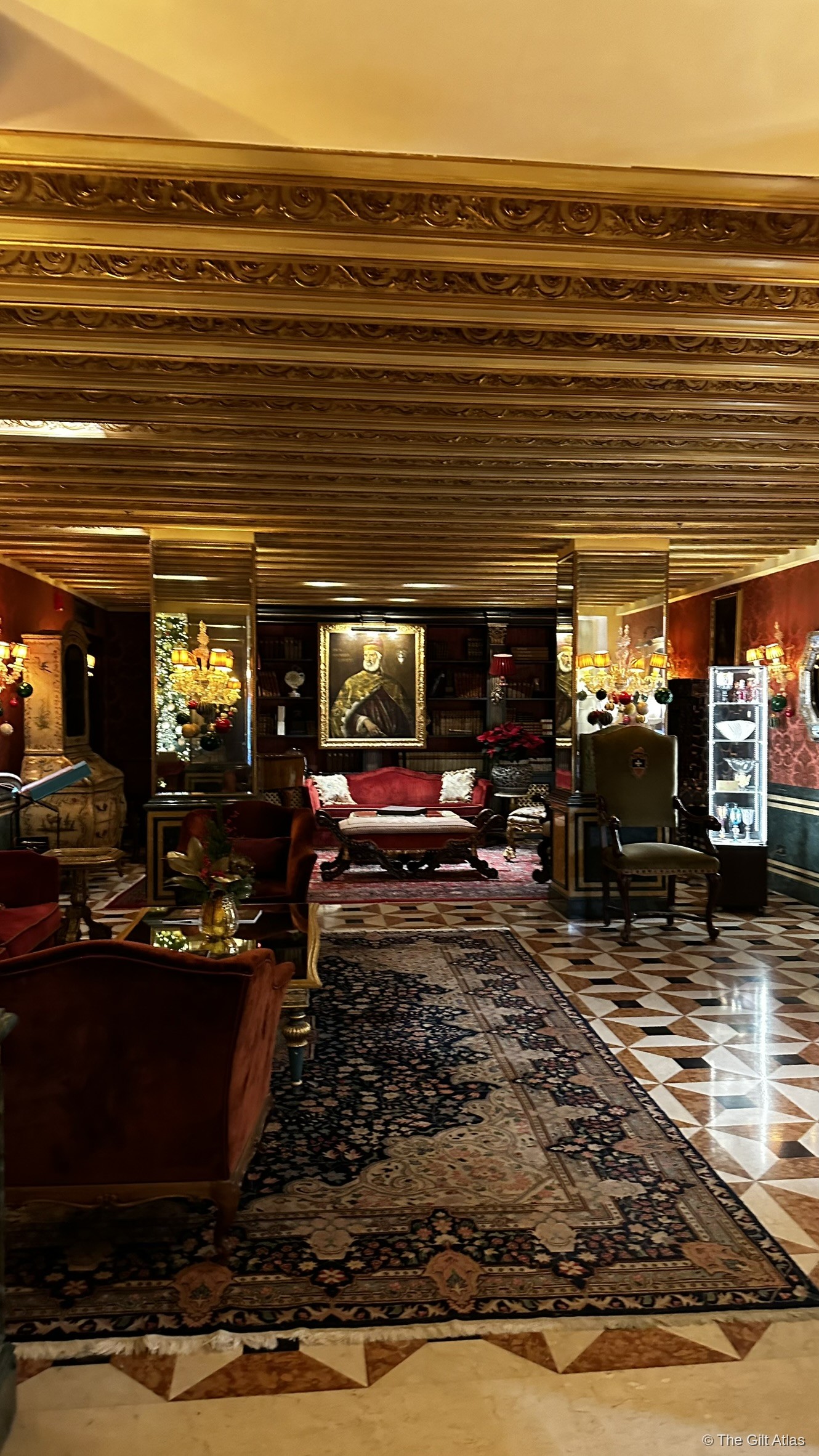 The Gritti Palace grand lobby — gold-coffered ceiling, red velvet furniture, antique portrait, and geometric Venetian marble floors