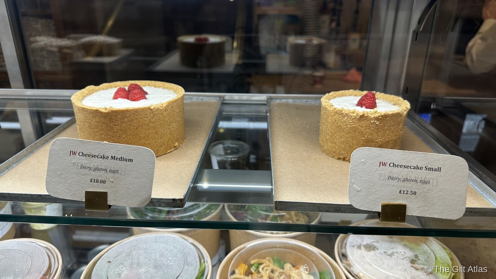 Two JW Cheesecakes on display in a glass case, labelled 'JW Cheesecake Medium £18' and 'JW Cheesecake Small £12.50'
