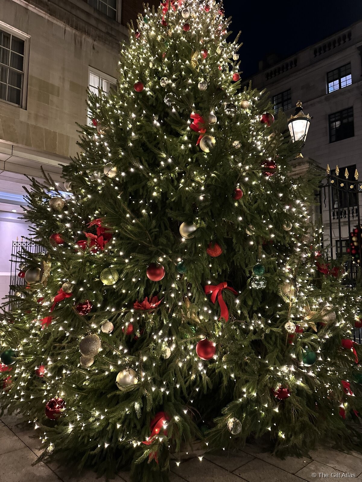 A towering illuminated Christmas tree outside the hotel at night, festooned with red and silver ornaments and warm white lights, with the Mayfair streetscape behind