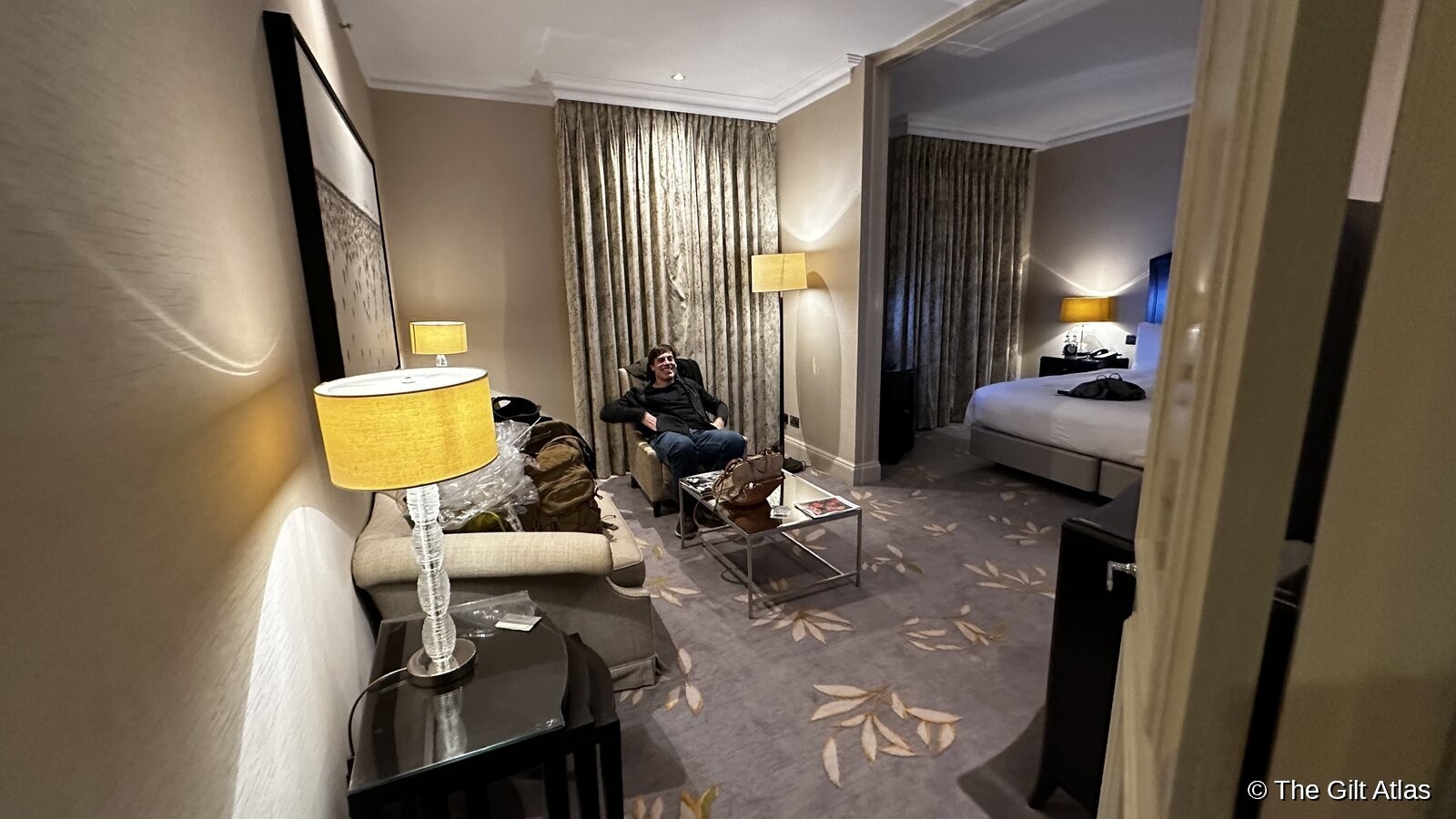 The sitting area of the Grosvenor House suite — warm lamps, floral carpet, velvet seating, and the bedroom visible through an archway beyond