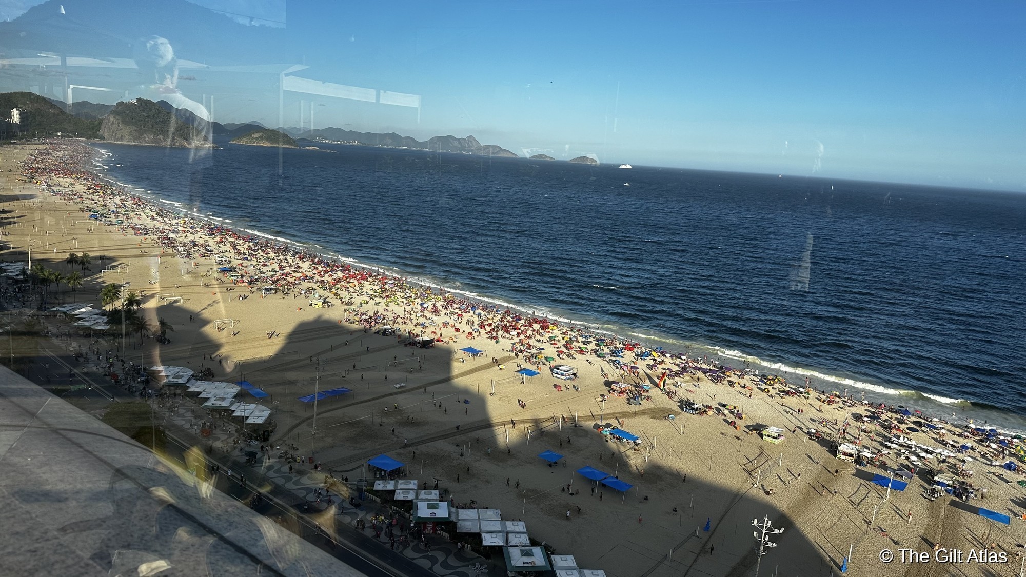 Wide-angle view of Copacabana beach from JW Marriott room — late afternoon long shadows, palm trees along the promenade, Dois Irmãos visible at far end