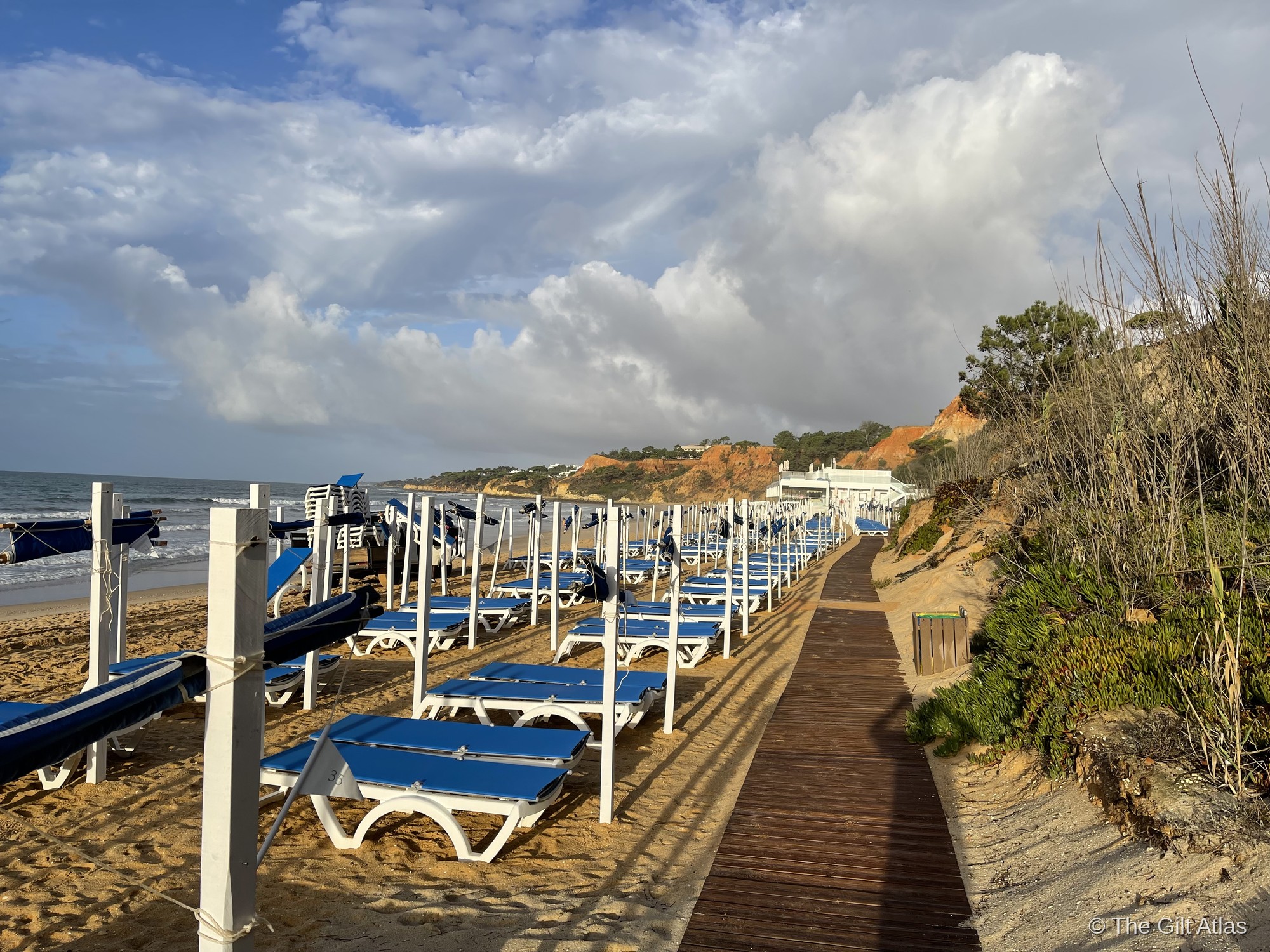 Blue sun loungers along the Pine Cliffs beach boardwalk at Praia da Falésia, with the ochre cliffs and pine trees behind and the Atlantic to the left