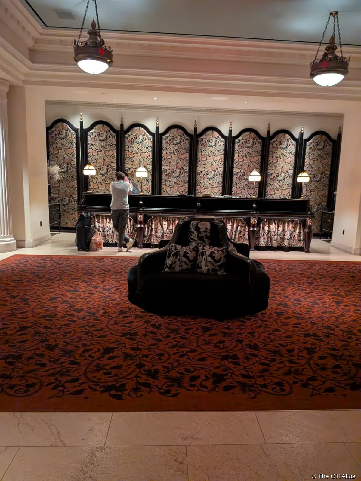 The Reserve Las Vegas check-in desk — ornate William Morris-style folding screen in jewel-toned florals behind the low reception counter, warm pendant lighting, marble floor, no queue