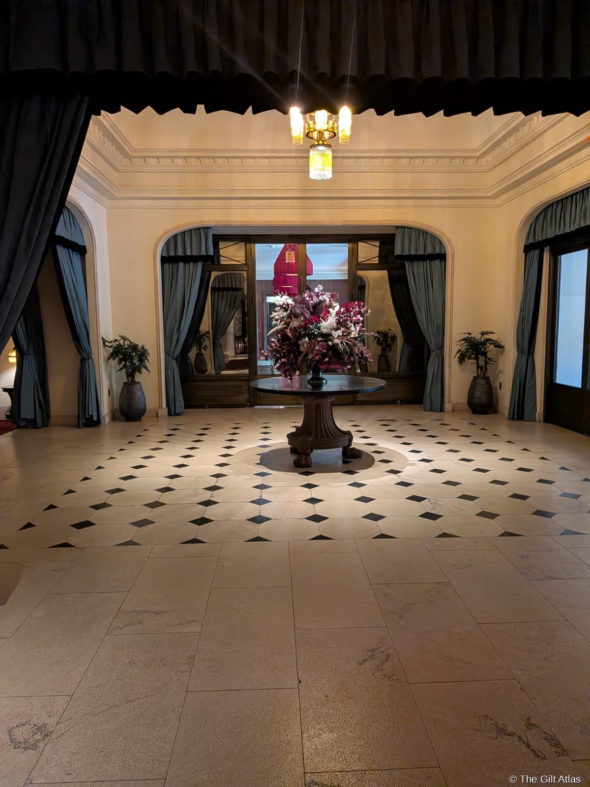 The Reserve Las Vegas entrance vestibule — marble tile floor with diamond inlay pattern, dramatic floral arrangement on central pedestal table, teal curtains framing the doorway to the lobby beyond