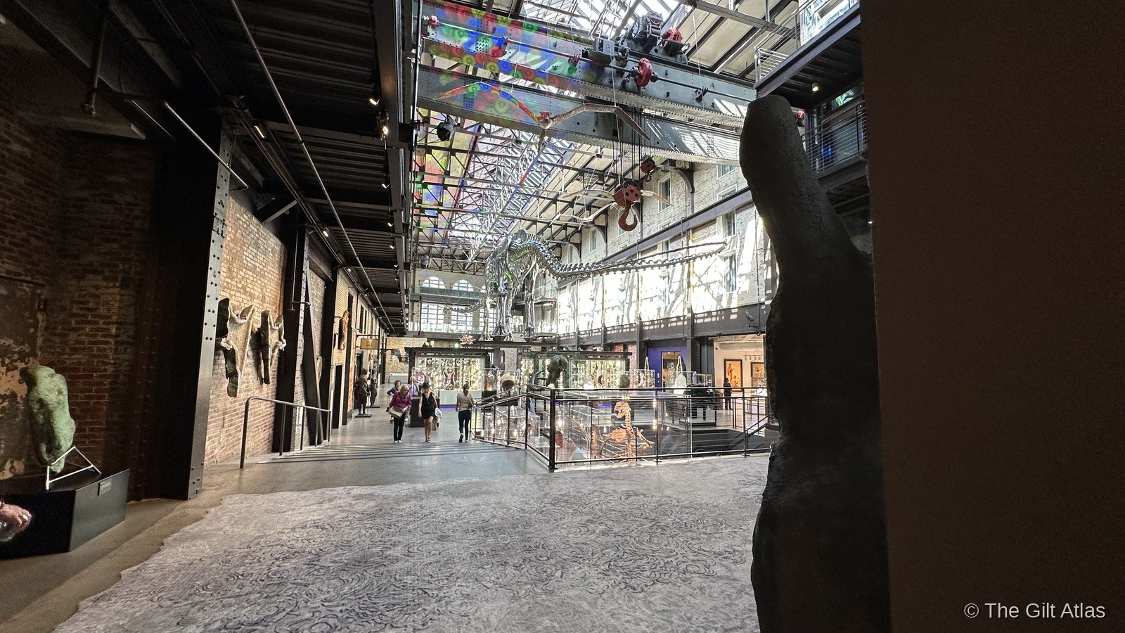 The Plant Riverside District main atrium — the soaring industrial brick hall with a glass skylight ceiling, hanging dinosaur skeleton, colorful LED installations, and the original power plant structure