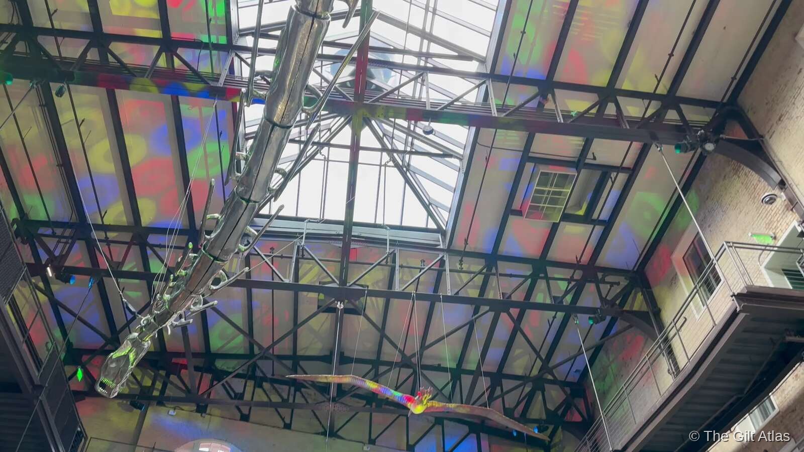 Looking up at the Plant Riverside District atrium ceiling — a full-scale dinosaur skeleton suspended in the air against the colorful LED-lit glass skylight and industrial steel framework