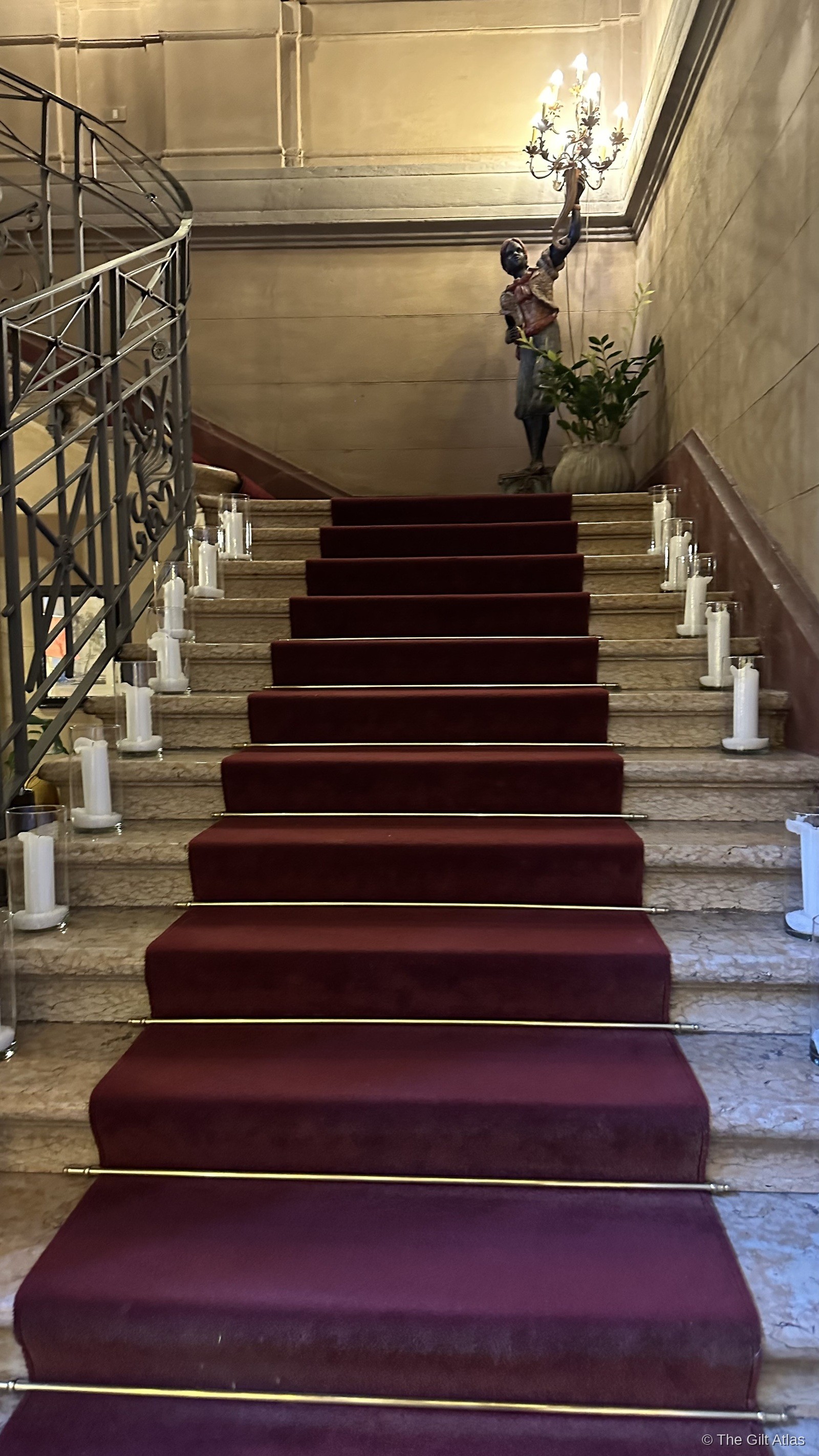 Hotel Terminus grand staircase — red carpet, marble steps, candles lining each side, bronze figure holding a candelabra above