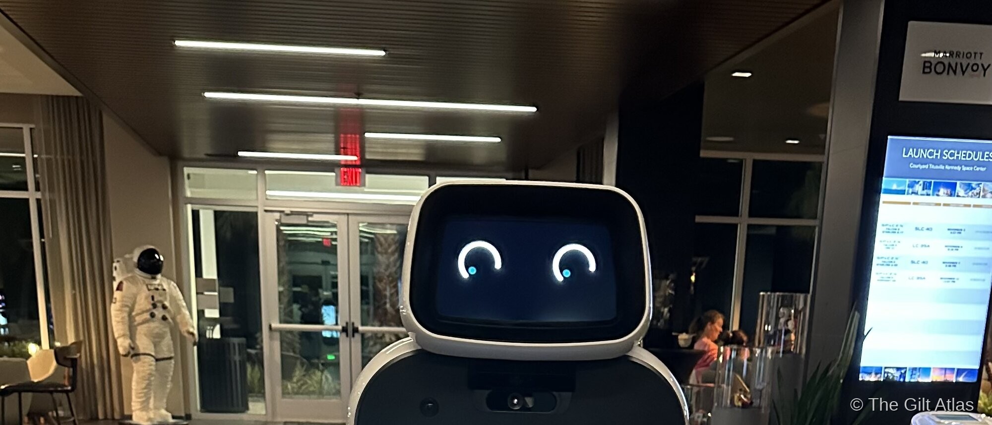 The LG CLOi robot assistant in the lobby of the Courtyard Titusville Kennedy Space Center — astronaut suit statue behind it, launch schedule board and a second CLOi unit visible in the background, Marriott Bonvoy signage above