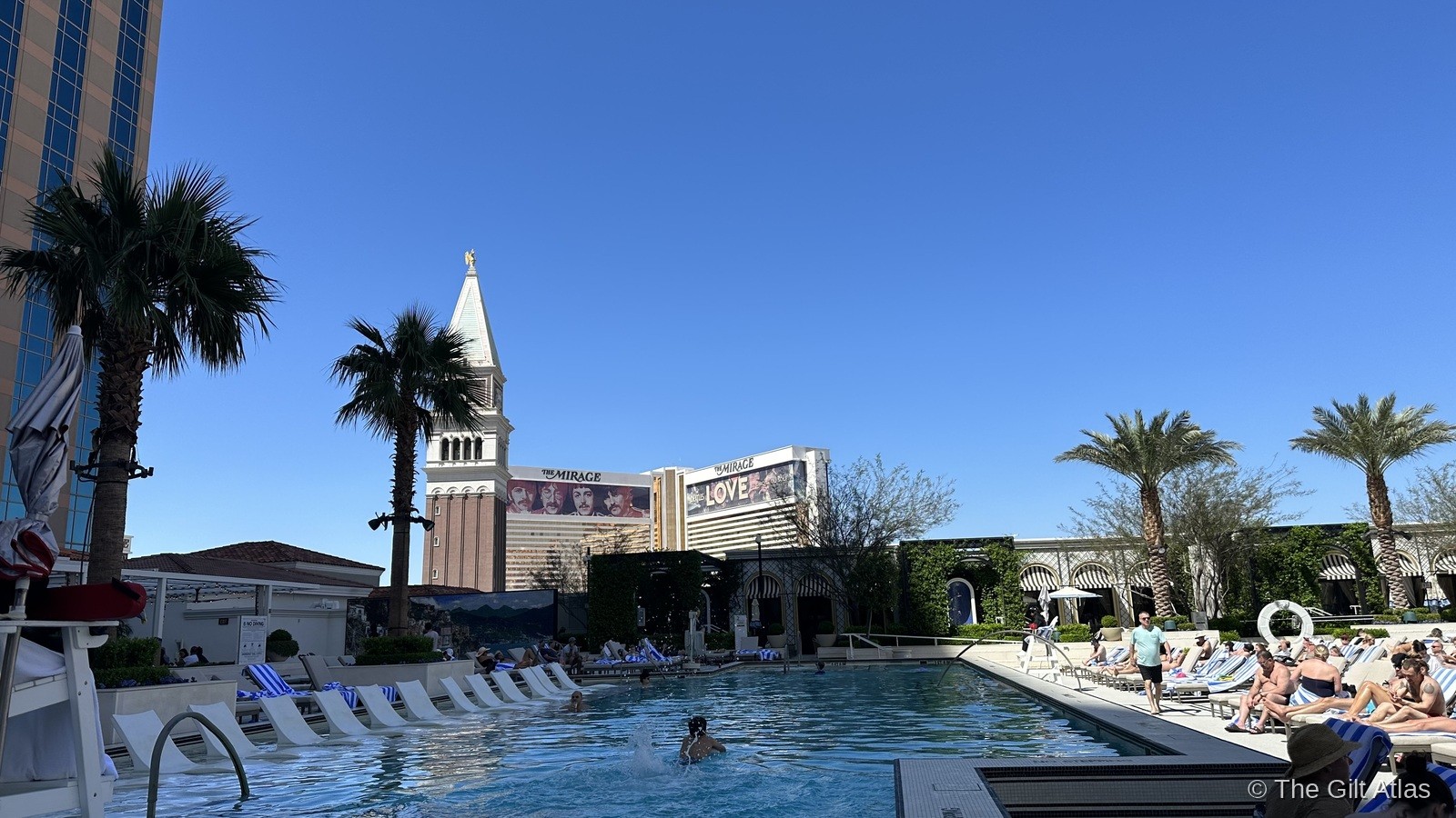 The Venetian pool deck — palm trees, lounge chairs, and the Campanile tower beyond with The Mirage in the background