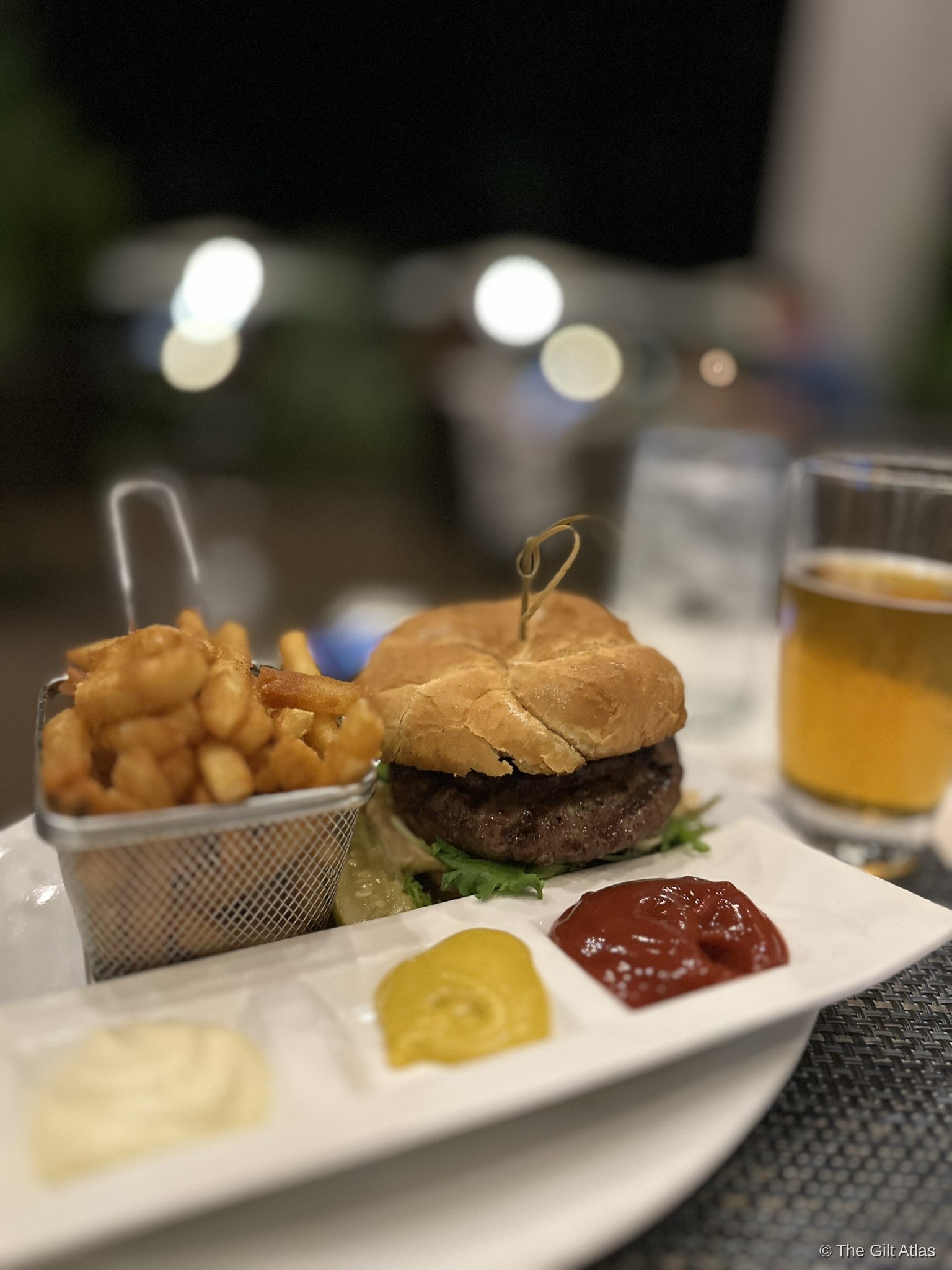A burger and fries with condiments and a beer at Lava Lava Beach Club, with string lights visible in the background