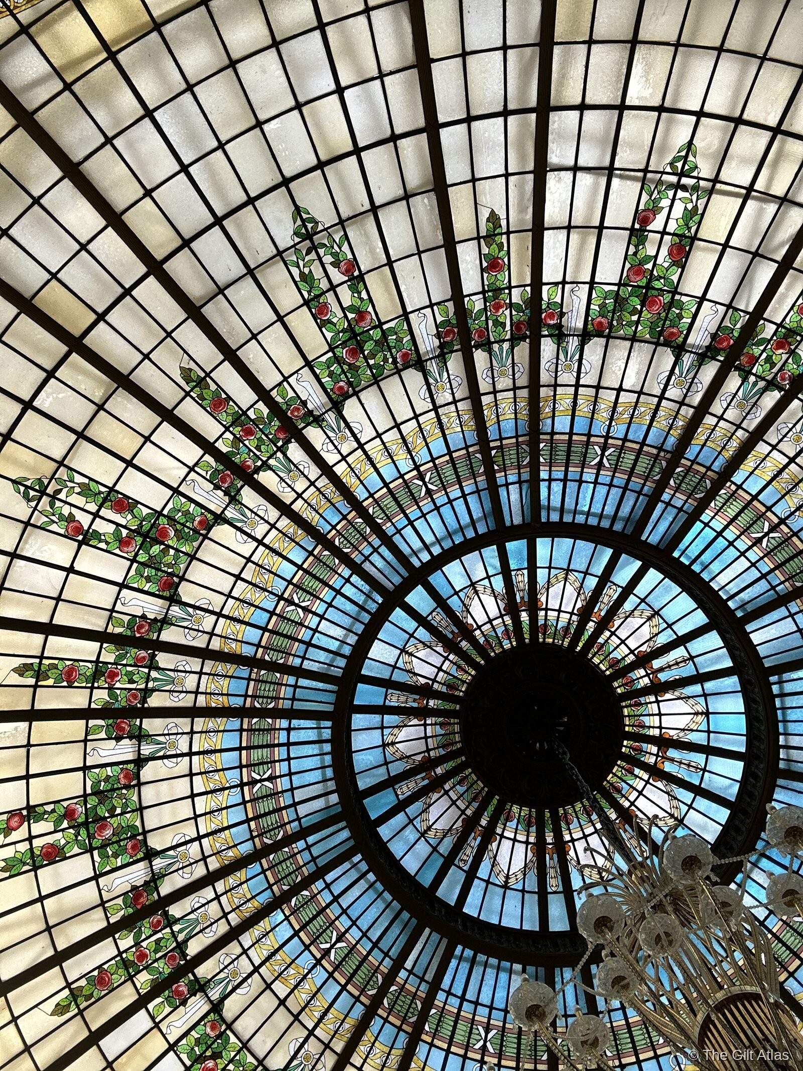 Close-up looking straight up at the Art Nouveau stained glass dome — concentric rings of roses, leaves and geometric patterns in green, red and cream, with a crystal chandelier at the edge