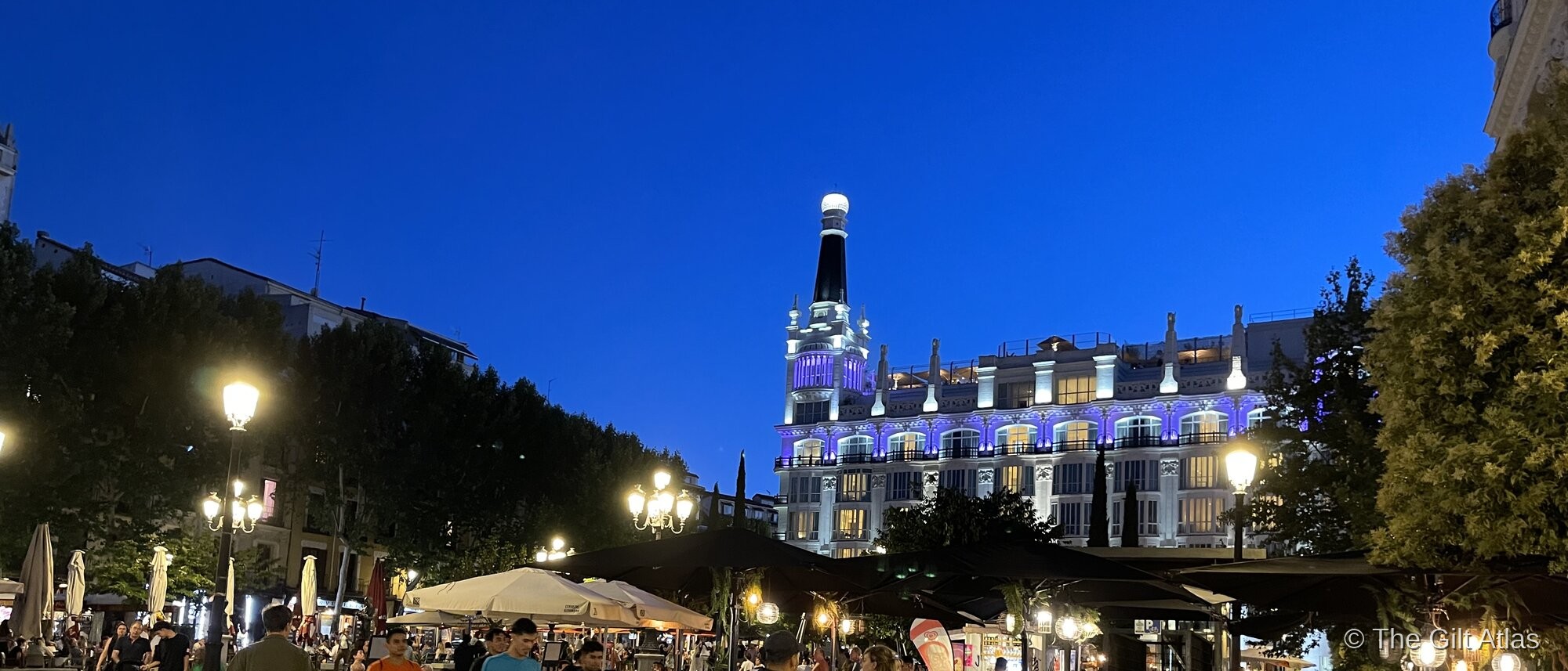 The Westin Palace Madrid illuminated at blue hour, the ornate white Belle Époque facade and distinctive black dome tower glowing against a deep blue sky, the busy Plaza de las Cortes in the foreground