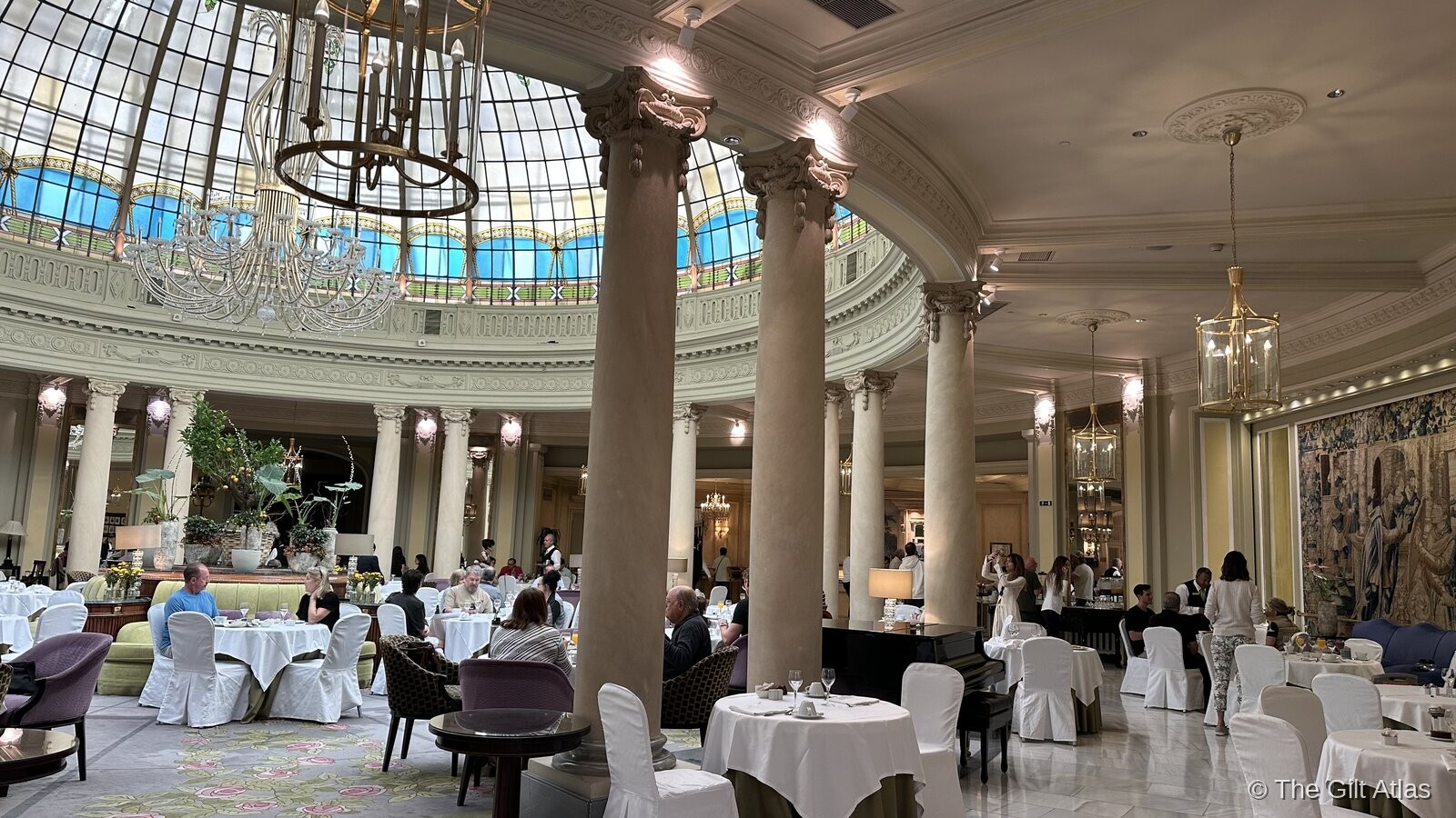 La Cúpula rotunda at the Westin Palace Madrid — Corinthian columns, stained glass dome overhead, white-linen tables filled with diners beneath chandeliers and a tapestry wall