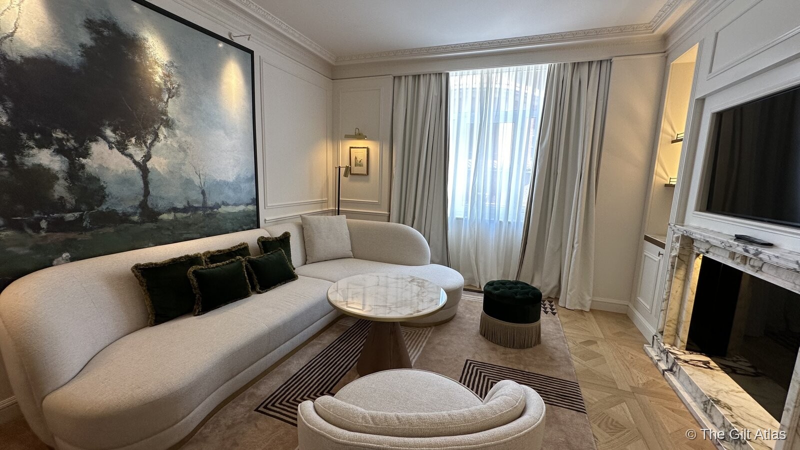 The renovated suite sitting room — curved cream sofa with emerald green velvet cushions, marble coffee table, large oil painting of trees, marble fireplace, parquet floors